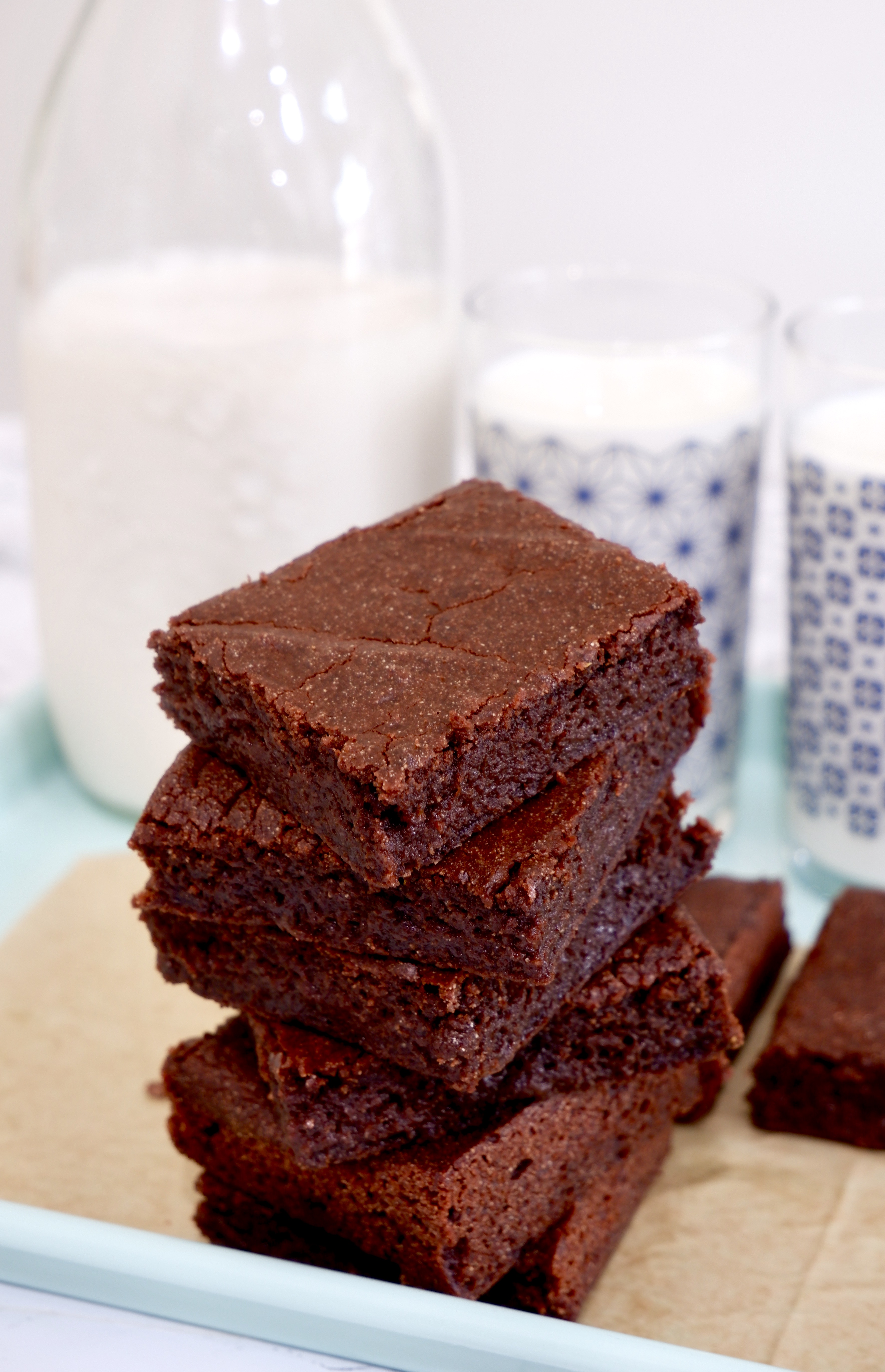 Six square mochi brownies stacked on top of each other; a glass bottle and two glasses of milk sit behind the stack of brownies; everything is on brown parchment paper and a mint colored tray