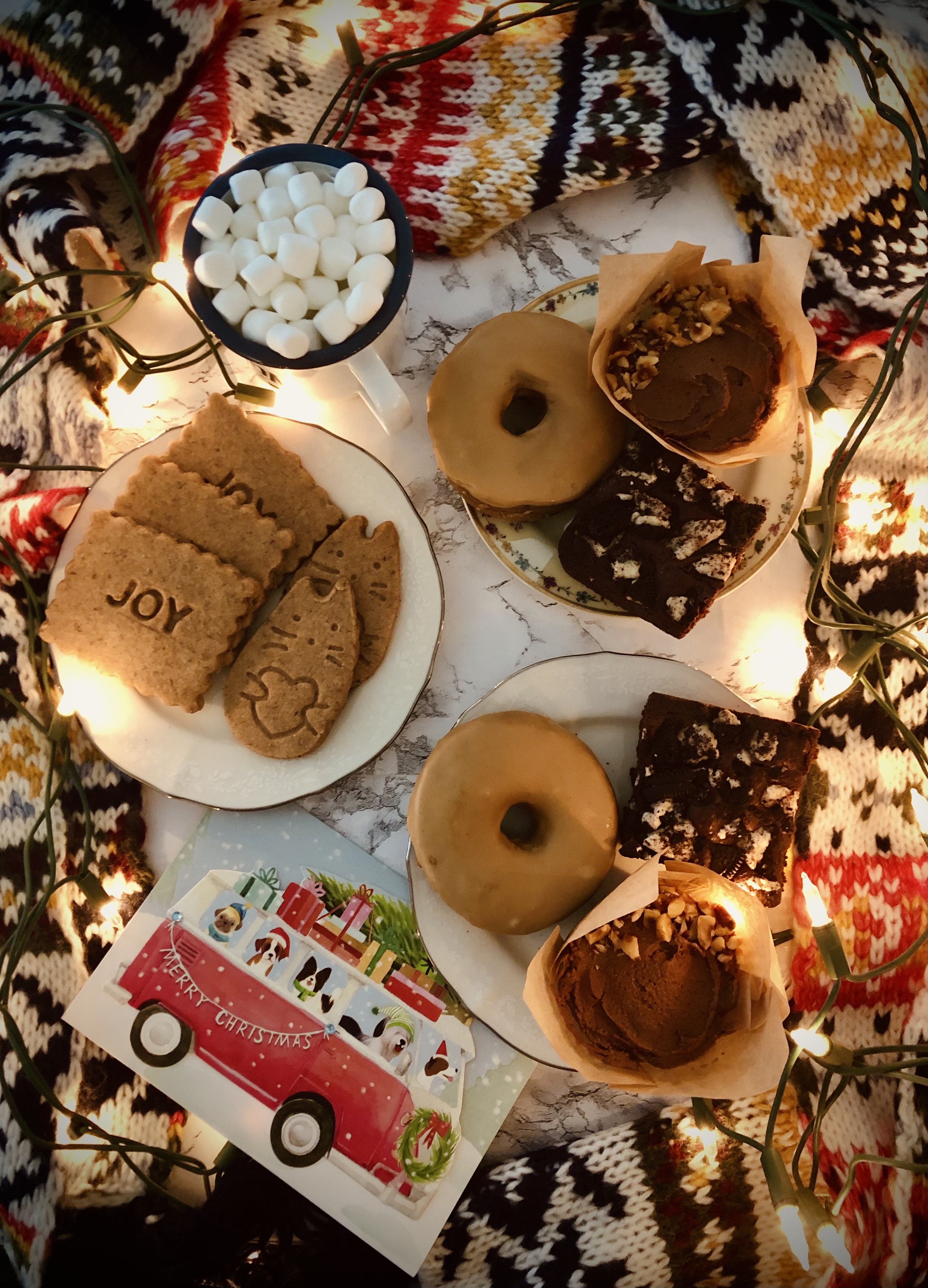 Overhead view of plates of cookies, donuts, brownies, and cupcakes, surrounded by a yarn knit scarf and holiday lights; dog holiday card and cup of marshmallows